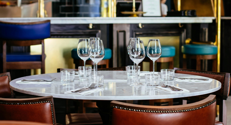 Mesa redonda de mármore posta para quatro em restaurante escuro, com guardanapos teal, taças e velas acesas, mesas ao fundo desfocadas