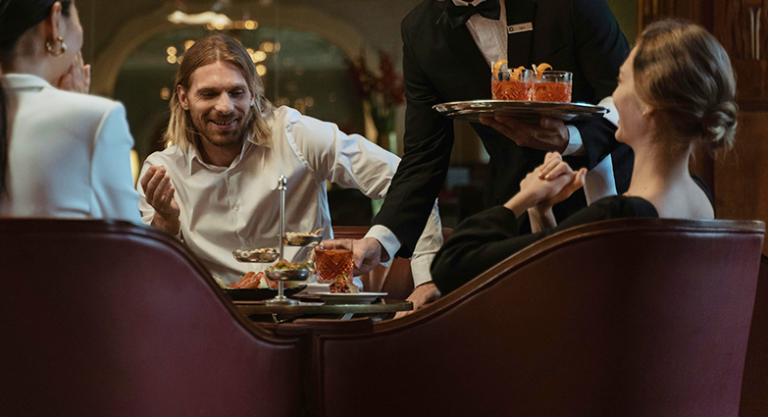 Garçom servindo drinques a grupo de Convidados em restaurante sofisticado com iluminação ambiente quente