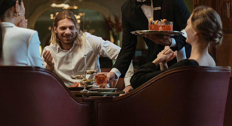 Garçom servindo drinques a grupo de Convidados em restaurante sofisticado com iluminação ambiente quente