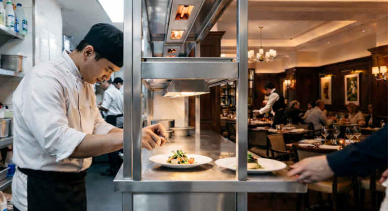Chef finalizando pratos no passe com salão elegante e iluminado visível ao fundo, ilustrando a separação entre bastidor funcional e ambiente de Convidados mostrando que estrutura traz de volta